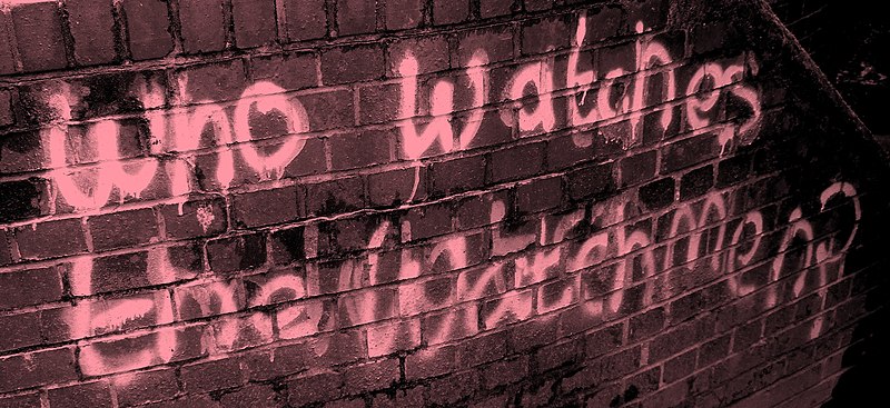 Un tag à la bombe écrivant "Who watches the watchers" (en française "Qui garde les gardiens?") sur un mur de brique.